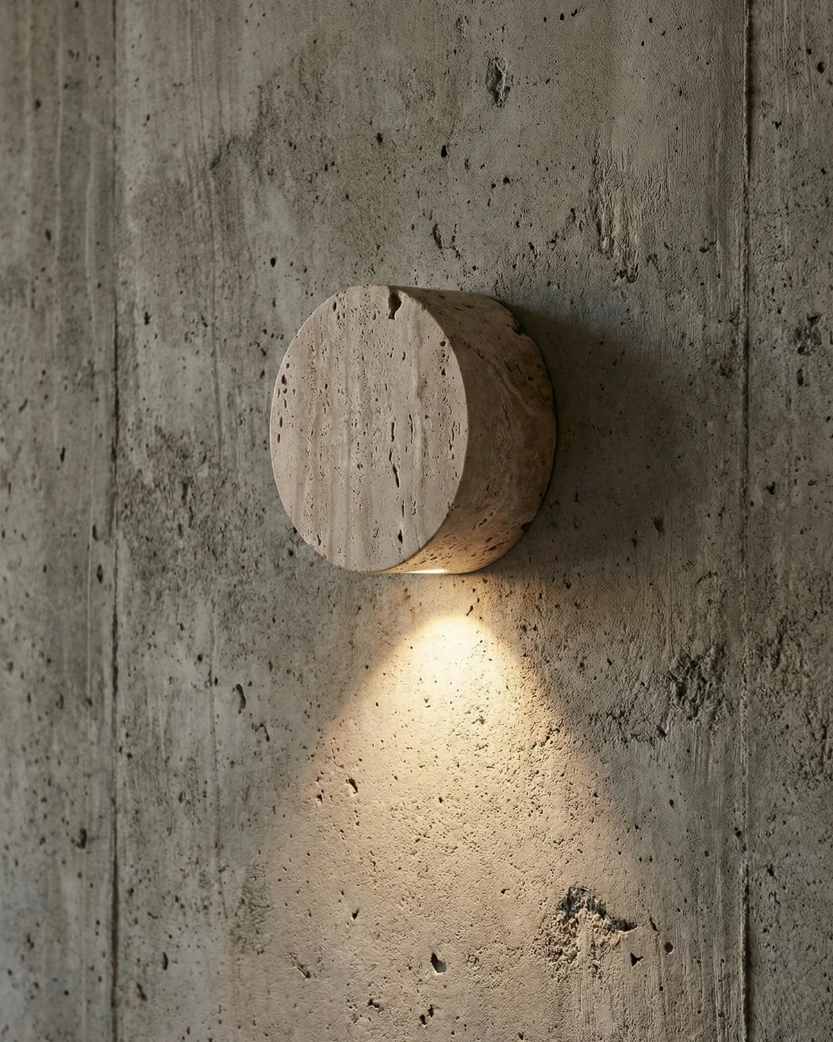 Travertine wall sconce VERONA No.01 lit on a raw concrete wall, travertine disc with light spilling below
