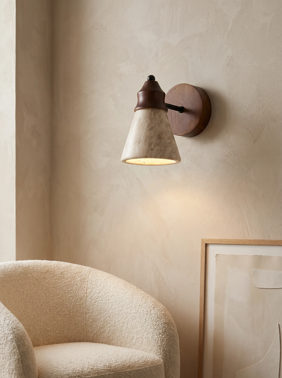 Travertine wall sconce CONA No.03 switched on above a cream boucle armchair and a leaning framed print