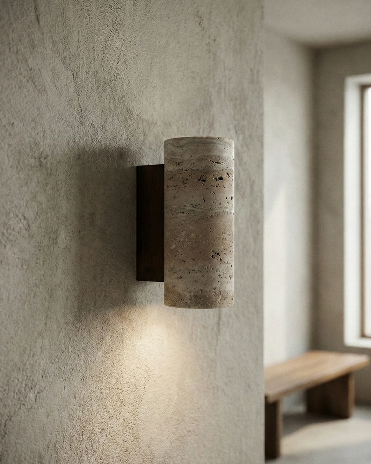 Travertine wall sconce GRANO No.02 lit on a plaster wall, casting light up and down toward a wooden bench
