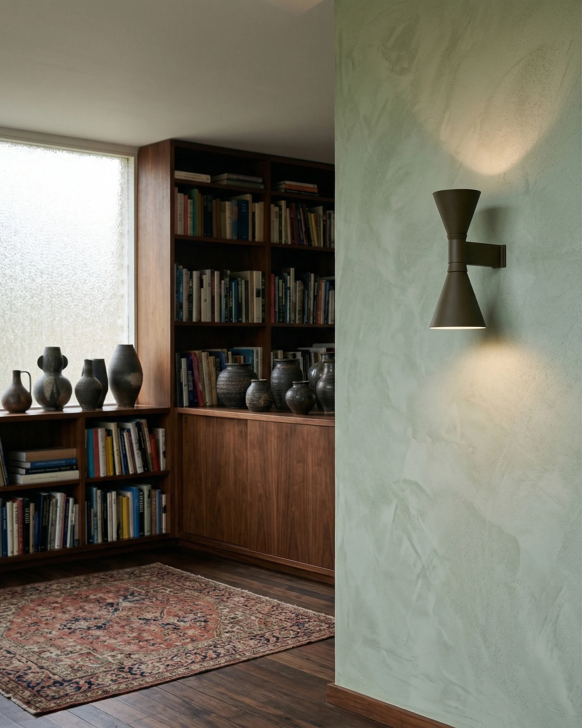 Up down wall sconce ECLISSI No.01 lit on a sage green plaster wall next to walnut bookshelves and a rug