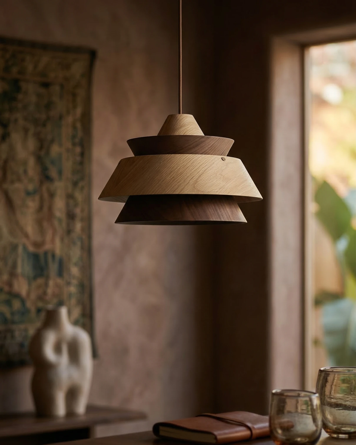 Wood pendant light BOSCO No.01 in light oak hanging above a table with glasses and a sculpture in front of a tapestry
