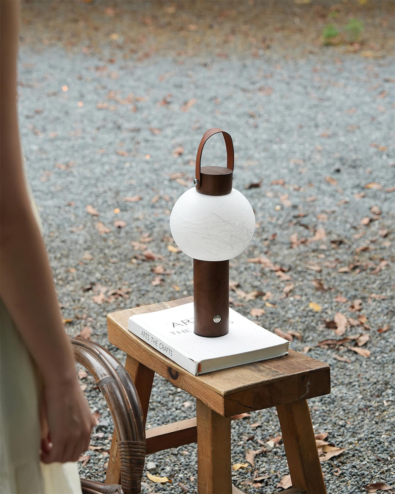 Wood table lamp NAPOLI No.01 lit on a small wooden stool on a gravel path outdoors, a hand reaching for it
