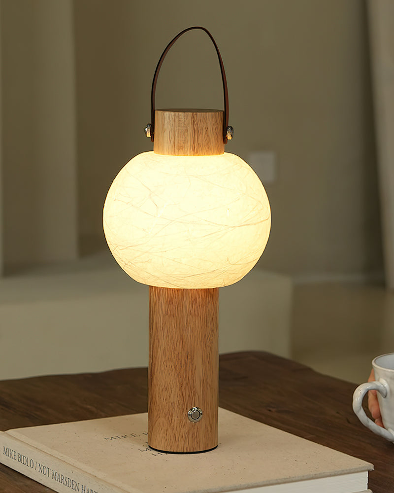 Wood table lamp NAPOLI No.01 in light oak switched on, standing on a stack of books with a mug beside it