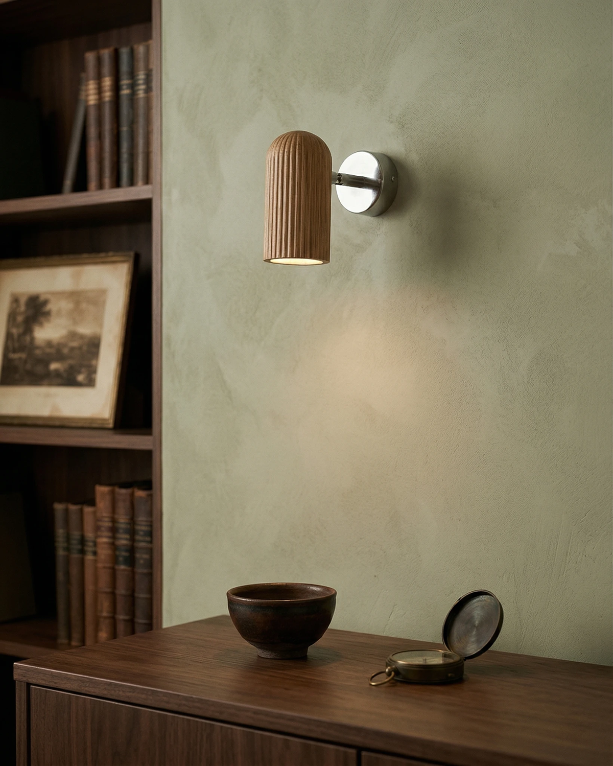 Wood wall sconce NOTA No.01 in walnut lit on a green wall above a wood cabinet beside tall bookshelves