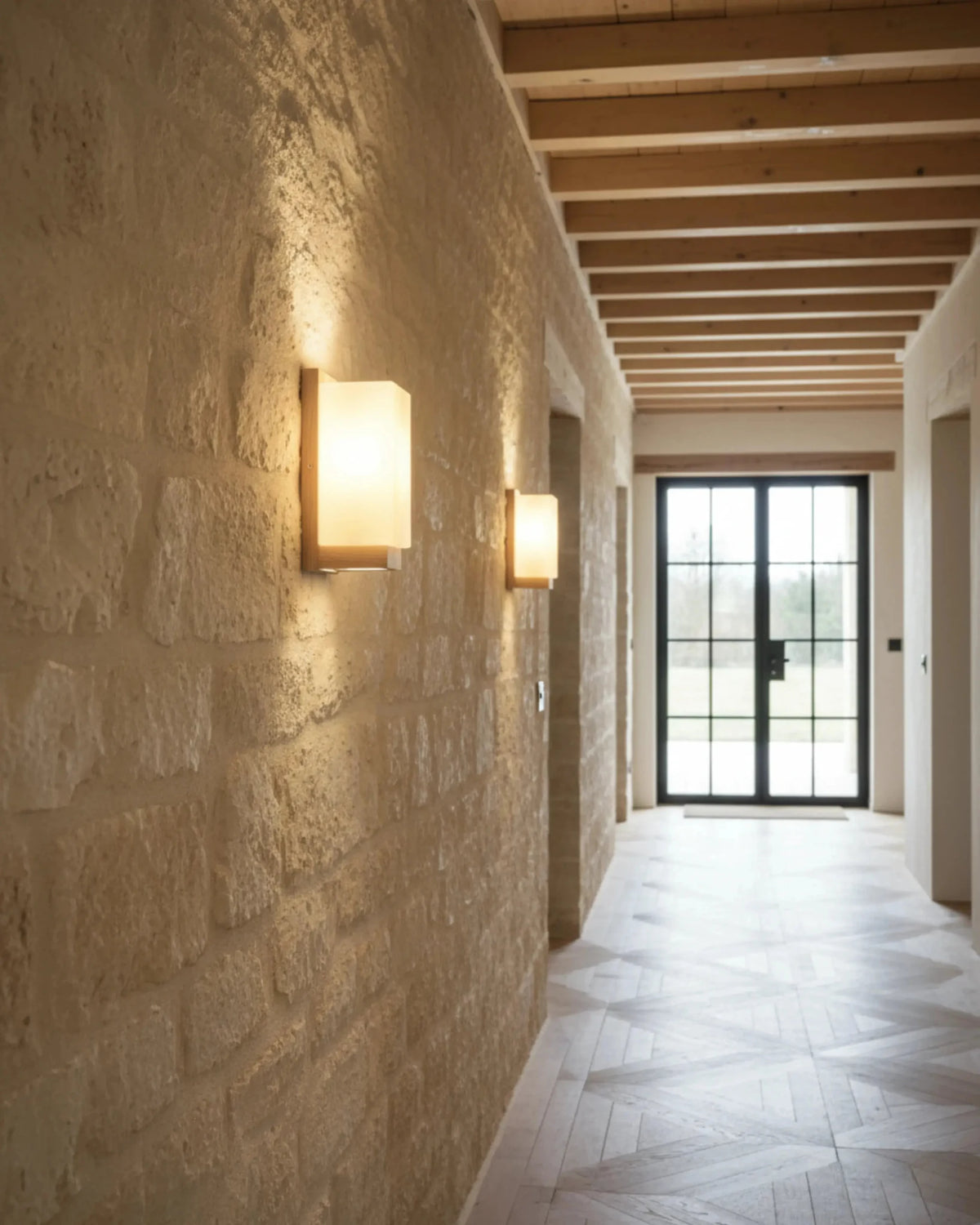 Two wood wall sconces MURO No.01 lit along a limestone corridor leading to a glass door