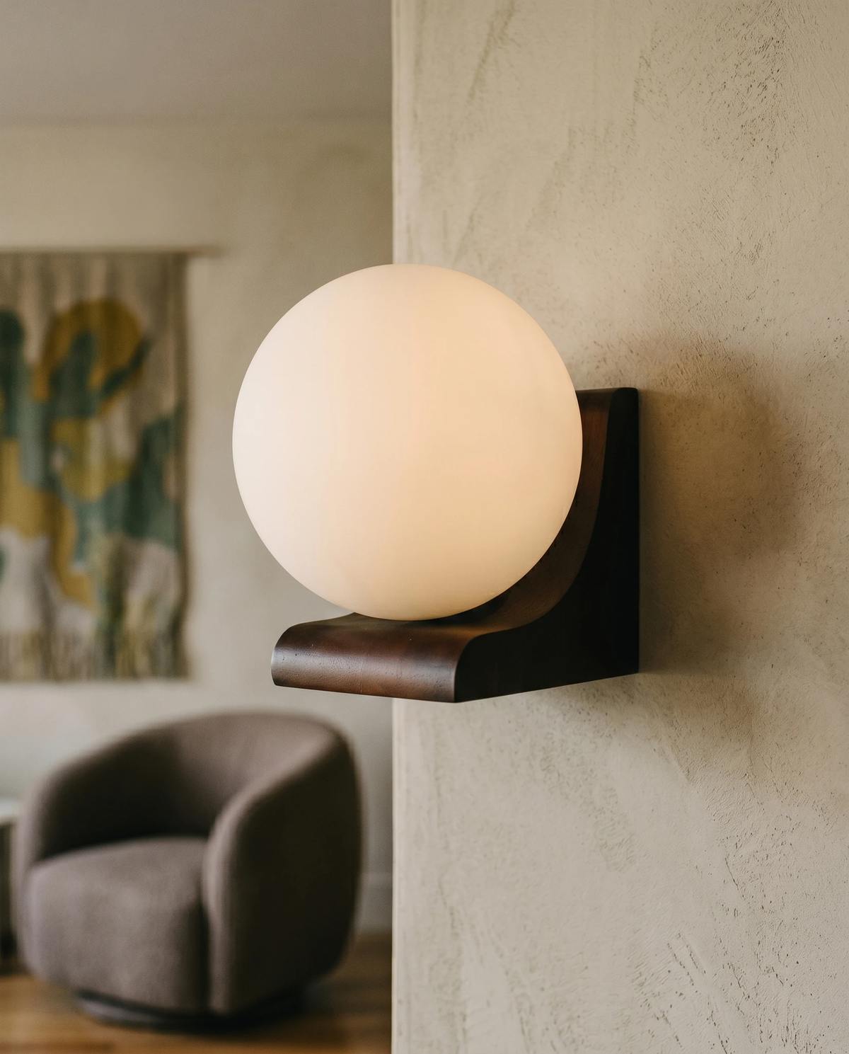 Wood wall sconce CURVA No.01 switched on beside a gray armchair and a patterned curtain on a plaster wall