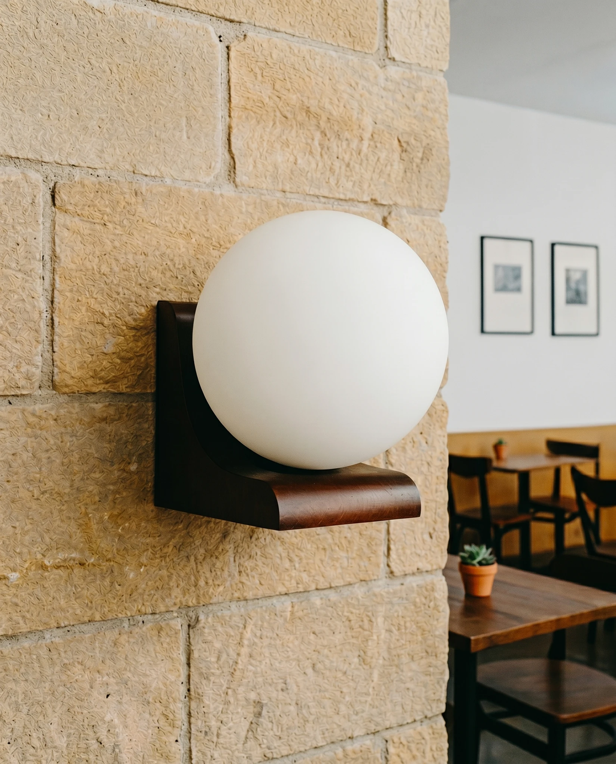 Wood wall sconce CURVA No.01 with an unlit frosted globe on a sandstone wall, café tables and chairs behind