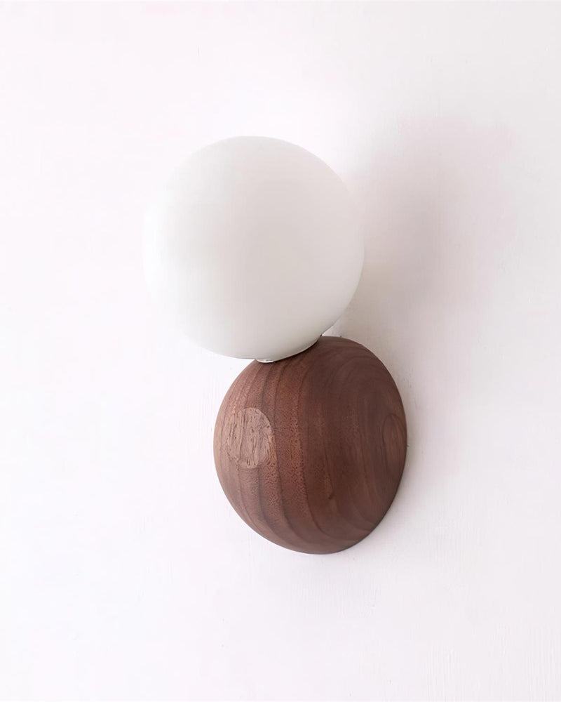 Wood wall sconce ALBE No.01 viewed straight on, matte opal globe above a walnut pebble base, switched off