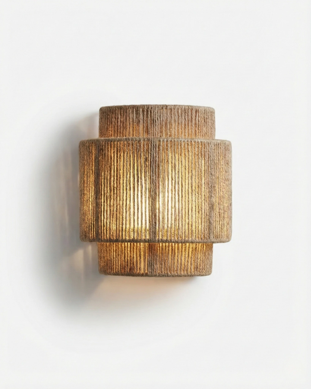 Woven wall sconce RAVENNA No.01 in Natural Jute, tiered rope-wrapped shade lit from within on a plain wall